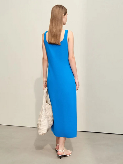 A woman facing away wearing a simple, minimalist, sleeveless midi dresses with a v-cut out in the neckline in blue. She has onn white strappy heels and a white bag to finish the look.