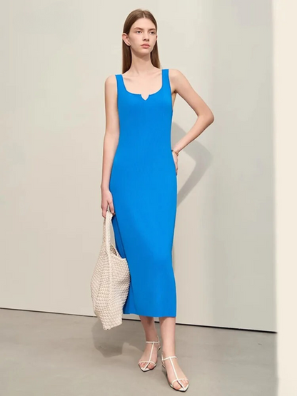 A woman wearing a plain, minimalist, sleeveless midi dresses with a v-cut out in the neckline in blue. She has on a pair of white strappy heels and a white bag to complete the look.