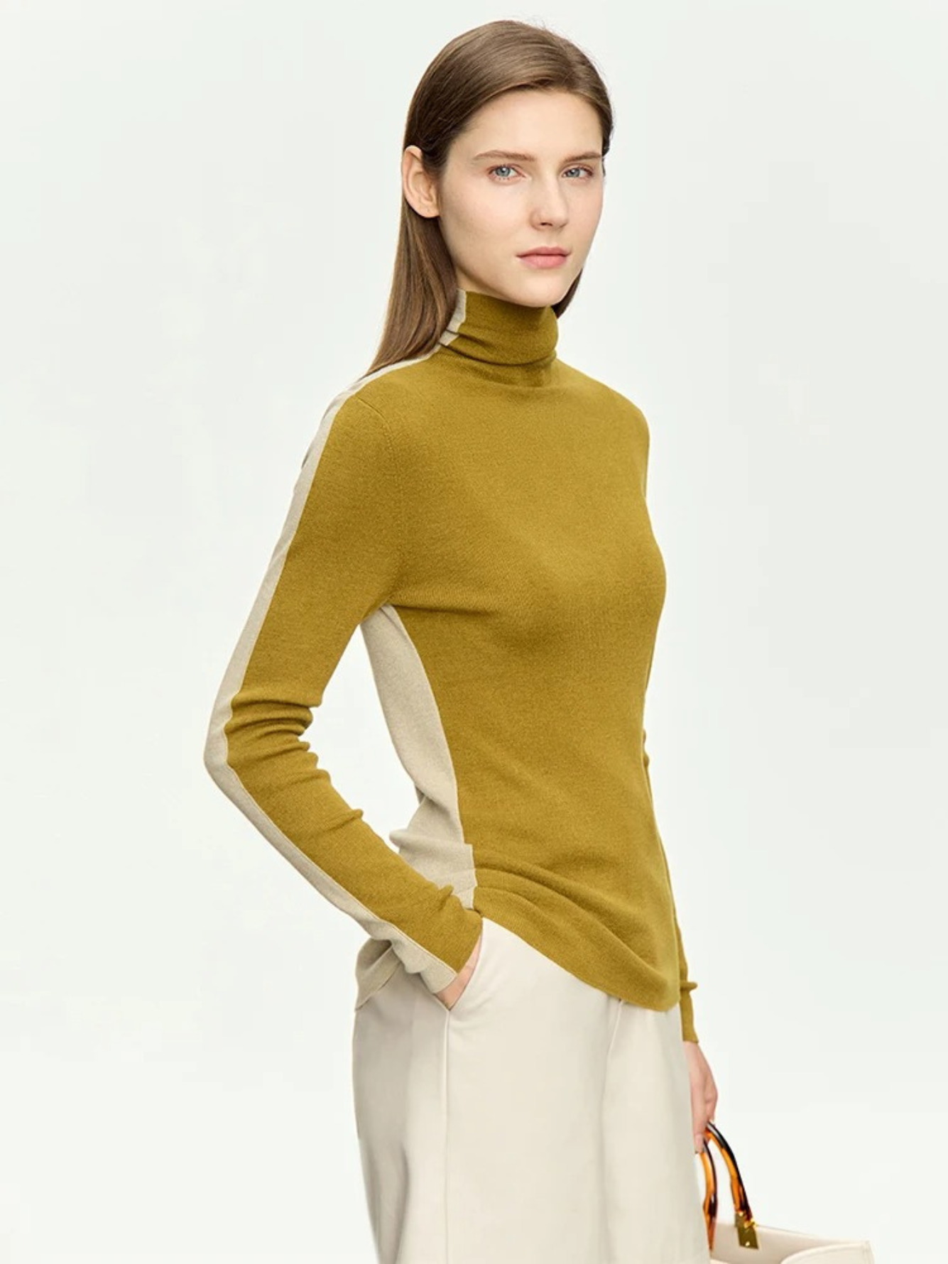 A woman wearing a mock neck, dual toned sweater in light yellow and mustard with a pair of cream colored high waisted pants with one hand in her pant pocket.